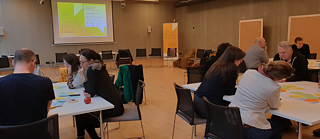 Conference or meeting room with participants seated at tables, facing a large screen displaying a presentation slide with text and graphics. The room includes posters or boards on the walls and chairs arranged in rows. Attendees appear to be engaged in discussions or activities related to the presentation