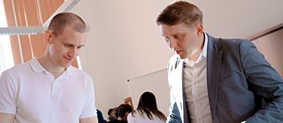 Zwei Personen betrachten gemeinsam etwas in einem hellen Büro- oder Schulungsraum; im Hintergrund sind weitere Personen und ein Flipchart zu sehen.