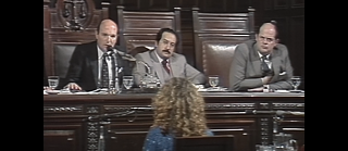 Drei Männer sitzen hinter einem großen Holztisch in einem Gerichtssaal. Ihre Gesichter sind unkenntlich gemacht. Vor ihnen sitzt eine Frau mit lockigem Haar und spricht in ein Mikrofon.