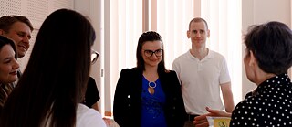 Mehrere Erwachsene stehen in einem Seminarraum im Gespräch. Im Hintergrund sind ein Whiteboard und ein Bildschirm zu sehen.