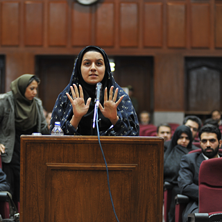 Eine Frau in dunkler Robe steht hinter einem hölzernen Rednerpult und spricht in ein Mikrofon. Beide Hände sind erhoben, die Handflächen zeigen nach vorne. Im Hintergrund sitzen mehrere Personen auf roten Stühlen.