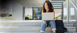 Studentka sedí na schodech před univerzitou. Na kolenou má notebook a s úsměvem hledí do dálky.