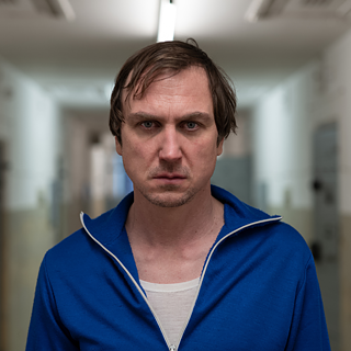 Mann in einem blauen Trainingsanzug steht in einem langen, hell erleuchteten Flur mit weißen Wänden und beigen unteren Wandabschnitten. Im Hintergrund sind mehrere Türen zu sehen.