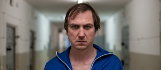 Ein Mann in einem blauen Trainingsanzug steht in einem langen, hell erleuchteten Flur mit weißen Wänden und beigen unteren Wandabschnitten. Im Hintergrund sind mehrere Türen zu sehen.