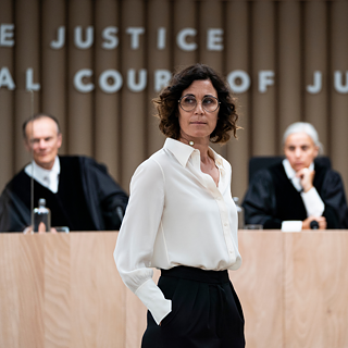 Eine Frau in weißem Hemd und schwarzer Hose steht vor einem Gerichtssaal. Im Hintergrund sitzen zwei Richter in schwarzen Roben an einem hölzernen Richtertisch. An der Wand hinter ihnen steht der Schriftzug „COURT OF JUSTICE“.