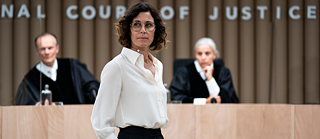 Eine Frau in weißem Hemd und schwarzer Hose steht vor einem Gerichtssaal. Im Hintergrund sitzen zwei Richter in schwarzen Roben an einem hölzernen Richtertisch. An der Wand hinter ihnen steht der Schriftzug „COURT OF JUSTICE“.