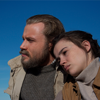 Dos personas están de pie muy cerca una de la otra frente a un cielo azul claro. Una persona lleva una chaqueta beige y un suéter gris, la otra persona lleva una chaqueta marrón y un suéter de cuello alto claro.