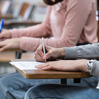 Studierende schreiben eine Prüfung in einem Klassenzimmer, Nahaufnahme der Hände mit Stift und Prüfungsbogen.