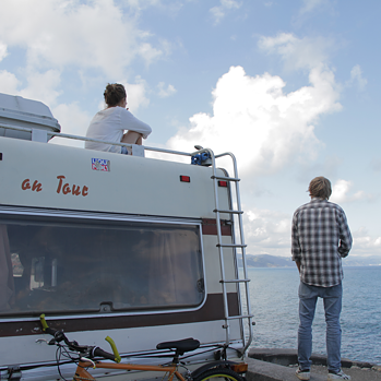 En gammel campingbus står med udsigt over havet, en ung kvinde sidder på taget, en ung mand står ved siden af køretøjet.