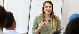 Üks naine seisab inimeste ees. Ta hoiab käes pliiatsit ja märkmikku, naeratab ja räägib. Taustal on valge tahvel. Tundub, nagu ta õpetaks või selgitaks midagi. 