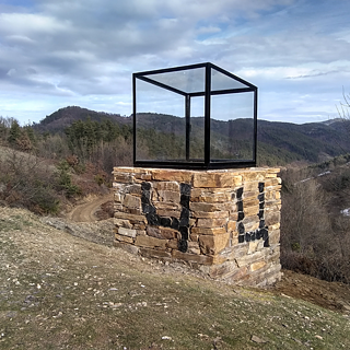 Skulptur von Terry Vreeburg mit gläsernem Würfel auf steinernem Sockel, der die kyrillischen Buchstaben ‚Ч‘ und ‚Ц‘ zeigt, platziert in bewaldeter Hügellandschaft unter teils bewölktem Himmel.