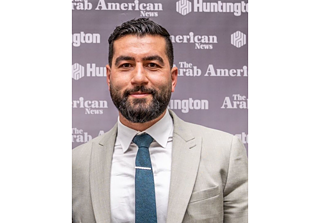 A bearded man slightly smiling in a light brown suit and navy blue tie
