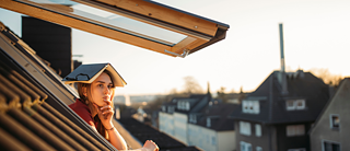 Eine Frau mit einem Buch auf dem Kopf blickt nachdenklich aus dem Dachfenster auf die Stadtlandschaft.