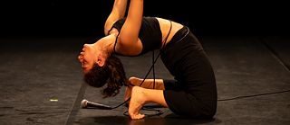 Eine Person führt auf der Bühne eine flexible Rückwärtsbewegung aus, bei der ihr Kopf den Boden berührt, und ein Mikrofon liegt daneben.