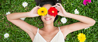 Eine Person liegt auf dem Rücken auf einer Wiese und hält zwei Blumen vor die Augen. Die linke Blume ist gelb, die rechte Blume ist rot. Um die Person herum liegen weitere bunte Blumen.
