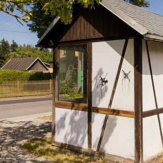 A half-timbered hut with white walls and dark wooden beams, used as a bus stop, stands on a sunny country road, surrounded by trees, bushes and other houses in the background.