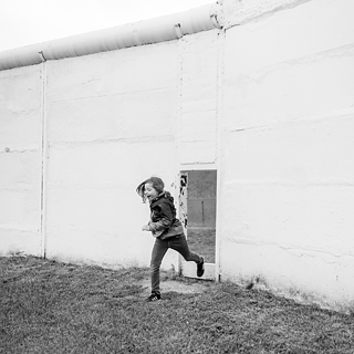 Черно-бяла фотография на човек, който тича през отвор в голяма бяла стена. Фигурата е заснета в движение, с единия крак във въздуха. Земята е покрита с трева, а стената има грапава текстура с видими линии. Сцената предава усещане за движение и динамика.