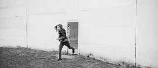 Черно-бяла фотография на човек, който тича през отвор в голяма бяла стена. Фигурата е заснета в движение, с единия крак във въздуха. Земята е покрита с трева, а стената има грапава текстура с видими линии. Сцената предава усещане за движение и динамика.