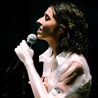 La cantante Camila Barrios canta frente a un micrófono. Tiene el cabello rizado, lleva una blusa blanca translúcida y una prótesis blanca y elegante en el brazo izquierdo. El fondo es oscuro, lo que la destaca como el centro de la imagen.