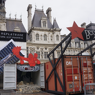 Kunstinstallation im Freien mit zwei Schiffscontainern. Ein blauer Container liegt seitlich und zeigt ein großformatiges Foto einer Person im Inneren. Ein orangefarbener Container steht aufrecht mit geöffneten Türen und dunklem Innenraum. Über den Containern befinden sich rote Sterne und abstrakte Formen. Ein schwarzes Banner mit kyrillischer Schrift hängt über dem orangefarbenen Container, daneben ein französisches Banner mit der Aufschrift: ‚PAS DE REPLÂTRAGE LA STRUCTURE EST POURRIE‘ (‚Kein Verputzen – die Struktur ist morsch‘). Im Hintergrund steht ein Gebäude mit klassizistischer Architektur.