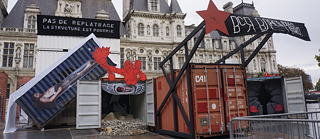 Kunstinstallation im Freien mit zwei Schiffscontainern. Ein blauer Container liegt seitlich und zeigt ein großformatiges Foto einer Person im Inneren. Ein orangefarbener Container steht aufrecht mit geöffneten Türen und dunklem Innenraum. Über den Containern befinden sich rote Sterne und abstrakte Formen. Ein schwarzes Banner mit kyrillischer Schrift hängt über dem orangefarbenen Container, daneben ein französisches Banner mit der Aufschrift: ‚PAS DE REPLÂTRAGE LA STRUCTURE EST POURRIE‘ (‚Kein Verputzen – die Struktur ist morsch‘). Im Hintergrund steht ein Gebäude mit klassizistischer Architektur.