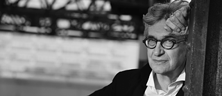 Schwarz-weiß-Fotografie einer Person im Anzug, die lässig an einer vertikalen Fläche lehnt, mit absichtlich unscharfem Gesicht, vor einem urban-industriellen Hintergrund aus Metallstrukturen und Gitterwand.