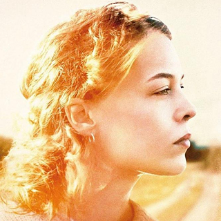 Person with curly, shoulder-length red-blond hair, photographed from the side, wearing a small earring.