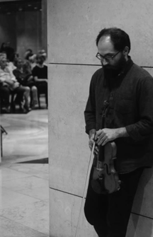 A fotografia a preto e branco mostra o artista David Magalhães Alves, encostado à parede, a olhar para o chão e com o violino na mão. Ele usa óculos e, ao fundo, à esquerda da parede, vê-se desfocada uma plateia sentada em cadeiras.