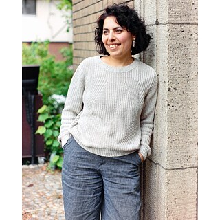 "امرأة تقف مبتسمة بجانب جدار حجري، ترتدي سترة صوفية فاتحة اللون وبنطال رمادي، وتبدو في وضع مريح في الهواء الطلق."