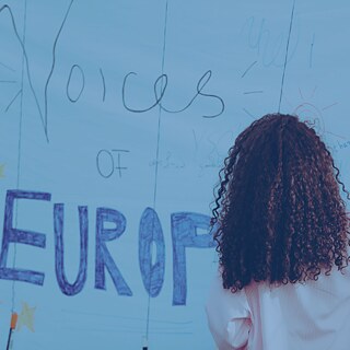 ier ist ein passender Alt-Text für das Bild: „Person mit lockigem Haar schreibt mit einem Stift auf eine große blaue Wand, die mit verschiedenen handgeschriebenen Nachrichten und Zeichnungen bedeckt ist. Im Hintergrund ist das Wort ‚EUROPE‘ in großen Buchstaben sichtbar.