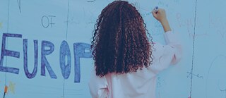 Person mit lockigem Haar schreibt mit einem Stift auf eine große blaue Wand, die mit verschiedenen handgeschriebenen Nachrichten und Zeichnungen bedeckt ist. Im Hintergrund ist das Wort ‚EUROPE‘ in großen Buchstaben sichtbar.