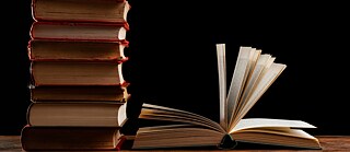Photo of a stack of books next to an open book laying on a table against a black background