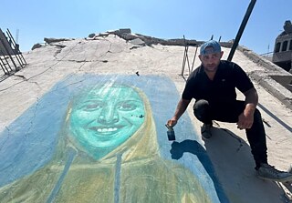 a man in a cap is posing besides his painting drawn on the debris of a bombed building.