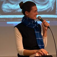 Person (Annabelle Hirsch) hält ein Mikrofon in der rechten Hand und ein Buch in der linken, trägt einen blauen Schal, im Hintergrund eine Projektion.
