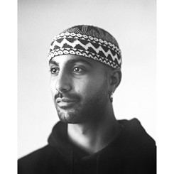 A man wearing a black and white patterned cap and an earring, looking to the right, with a soft light gray blurred background.