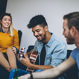 Das Bild zeigt vier Personen in entspannter Atmosphäre, die gemeinsam Zeit verbringen. Eine Person spielt eine akustische Gitarre, während die anderen aufmerksam zuhören und den Moment genießen. Die Szene vermittelt ein Gefühl von Geselligkeit, Freundschaft und gemeinsamer Freude an Musik.