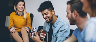 Das Bild zeigt vier Personen in entspannter Atmosphäre, die gemeinsam Zeit verbringen. Eine Person spielt eine akustische Gitarre, während die anderen aufmerksam zuhören und den Moment genießen. Die Szene vermittelt ein Gefühl von Geselligkeit, Freundschaft und gemeinsamer Freude an Musik.