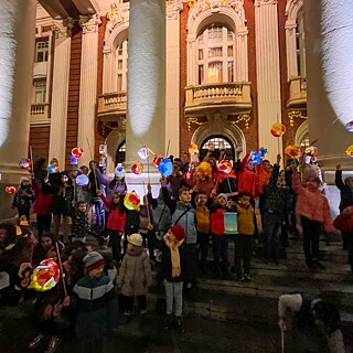 Kinder mit bunten, leuchtenden Laternen stehen auf den Stufen des Nationaltheaters „Ivan Vasow“ in Sofia. Die klassizistische Fassade mit ihren imposanten Säulen bildet die festliche Kulisse für den traditionellen Martinsumzug, der Licht und Gemeinschaft in die dunkle Jahreszeit bringt.