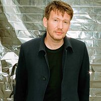 Person (Dmitrij Kapitelman) in dunkler Jacke steht vor silbrig glänzendem Hintergrund, trägt schwarzes Oberteil.