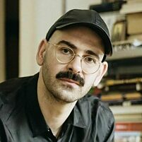 Person (Enrico Ippolito) in schwarzem Hemd und schwarzer Kappe sitzt vor einem Regal mit Büchern und Gegenständen.