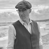 Schwarz-weiß-Foto einer Person (Ewald Arenz) am Strand, trägt Hemd, Weste und Schiebermütze, Meer im Hintergrund.