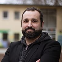 Person (Holger Marcks) im dunklen Mantel und Kapuzenpullover steht mit verschränkten Armen im Freien, unscharfer Hintergrund mit Gebäuden.