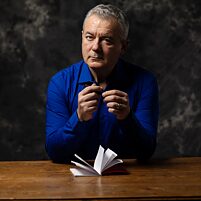 Person (Ilija Trojanow) in blauem Hemd sitzt an einem Holztisch mit aufgeschlagenem Notizbuch und Stift in der Hand, dunkler Hintergrund.