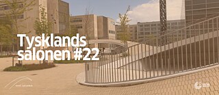 I baggrunden ses et billede af Karen Blixens Plads i København med forskellige bygninger og de karakteristiske bakkelignende forhøjninger, under hvilke der er cykelparkeringspladser.