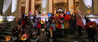 Kinder mit bunten, leuchtenden Laternen stehen auf den Stufen des Nationaltheaters „Ivan Vasov“ in Sofia. Die klassizistische Fassade mit ihren imposanten Säulen bildet die festliche Kulisse für den traditionellen Martinsumzug, der Licht und Gemeinschaft in die dunkle Jahreszeit bringt.