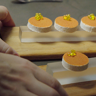 Primer plano de seis pequeños bocados redondos colocados sobre dos placas rectangulares de vidrio, que reposan sobre bases de madera. Los bocados tienen una base clara, una capa superior anaranjada y están decorados con pequeñas perlas amarillas.