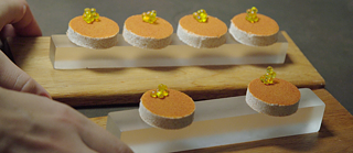 Primer plano de seis pequeños bocados redondos colocados sobre dos placas rectangulares de vidrio, que reposan sobre bases de madera. Los bocados tienen una base clara, una capa superior anaranjada y están decorados con pequeñas perlas amarillas.