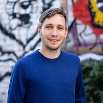 Person (Maik Fielitz) in blauem Pullover steht vor einer Wand mit bunter Graffiti-Kunst, im Hintergrund sind Pflanzen sichtbar.