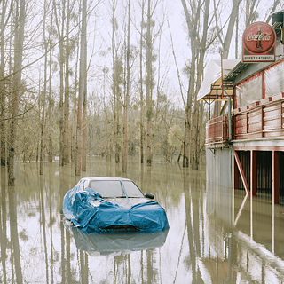  Une voiture enveloppée d’une bâche bleue est à moitié immergée, entourée d’arbres inondés et d’un bâtiment déserté portant une enseigne Coca-Cola délavée. La scène, à la fois calme et inquiétante, devient une métaphore de l’immobilité, de la fragilité et de la puissance de la nature. 