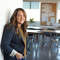 Persona con cabello largo y ondulado, vestida con chaqueta oscura y camiseta clara, apoyada contra una pared en un aula luminosa. Al fondo se observan varias mesas y sillas, una gran ventana, una pizarra de corcho con papeles y algunas plantas.